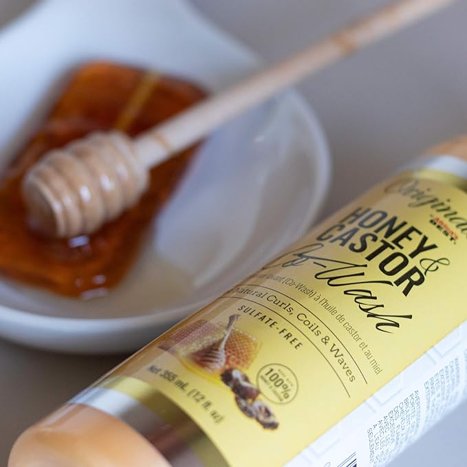 Yellow-labeled bottle reading Originals Honey & Castor Co-Wash, for Natural Curls, Coils & Waves, Sulfate-Free, Net 355 mL (12 fl oz), lying beside a small white dish of honey with a honey dipper.