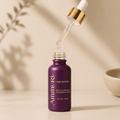 Purple 30ml dropper bottle labeled Ammuri Age Reverse, Botox Alternative, with gold-capped glass dropper releasing a drop of serum, beside a small bowl on a beige backdrop
