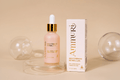 Pink glass dropper bottle with a gold collar labeled "Ammuri High Strength Retinol 3.5% Anti-Ageing Formula, 30ml", beside its matching white box, displayed on a clear glass pedestal with glass sphere props on a beige backdrop.